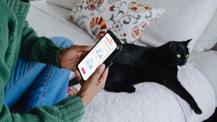 Een vrouw checkt op haar telefoon de Royal Canin Voedingswijzer terwijl ze op bed ligt met een zwarte kat. 